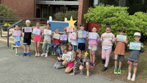 Fifteen students standing in front of Schofield School each holding a colorful sheet of paper with one letter on it spelling out ¡Bienvenidos!