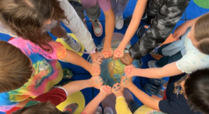 Ten students standing in a circle on a colorful rug with a globe in the center. The students hands meet in the middle, each student has a smiley face drawn on their hand in various colors