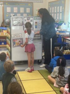 A student using the whiteboard with the help of a teacher while other students watch from the colorful rug
